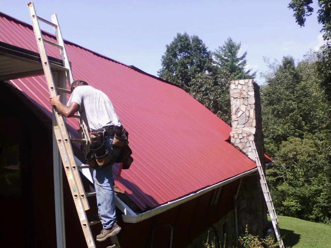 Roofer on a ladder beside a red metal roof and stone chimney — Soffit & Fascia Repair by Roofing Brothers in Matawan